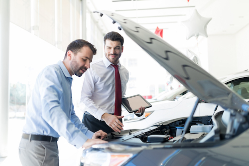 Smiling dealer holding digital tablet while looking at client analyzing car engine